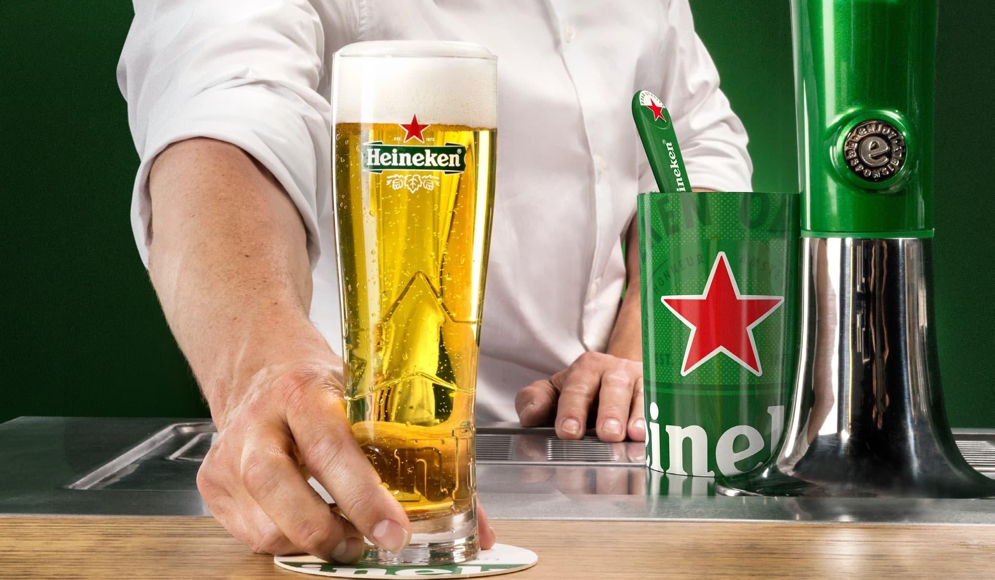 A bartender in a white shirt serves a freshly poured Heineken beer in a tall branded glass.